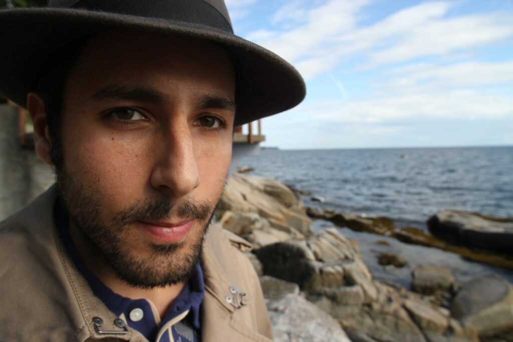 Colour photo of head and shoulders of Carmen, who has a beard and is wearing a hat with a brim, out of focus in the background are rocks and a body of water