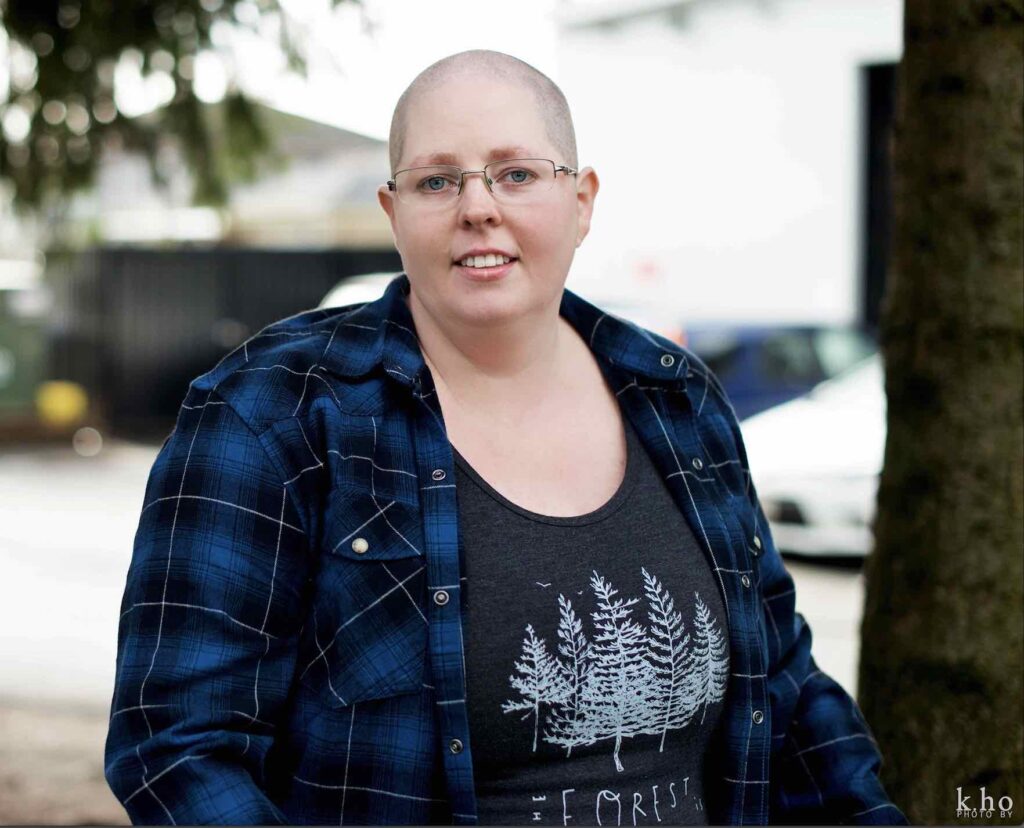 Colour photo of Heather with shaven head, wearing glasses, standing facing the camera, wearing a blue plaid shirt open over a black t-shirt with white trees on the front, they are outside under a tree 