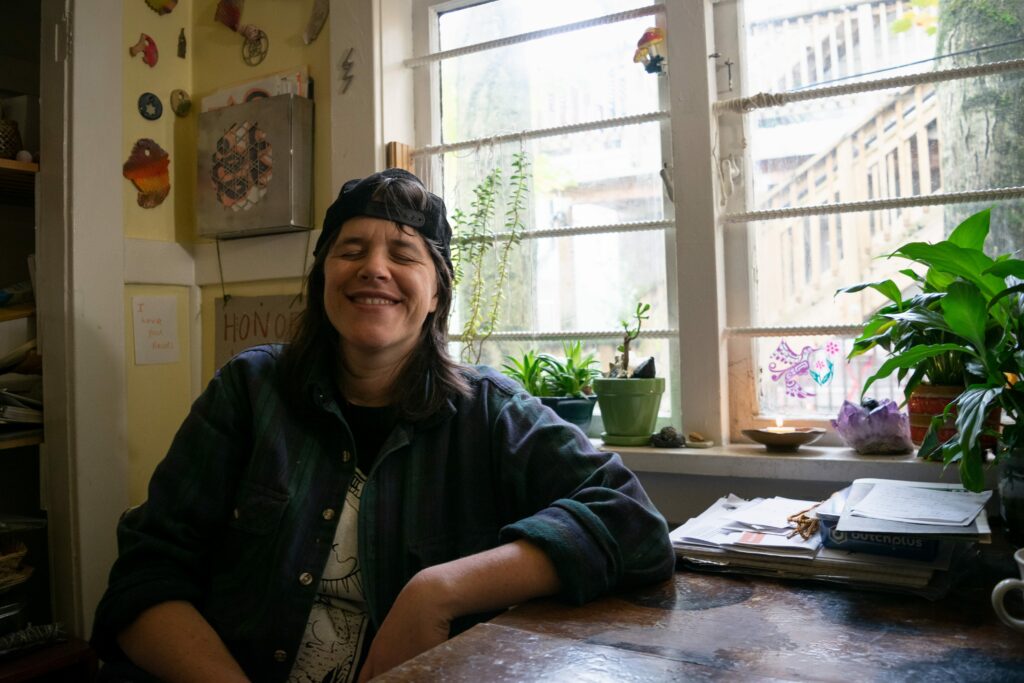 Colour photo of Heidi smiling, their eyes closed, sitting in front of a window, their left elbow on a table beside them, they have a dark shirt and dark baseball cap backwards on their head