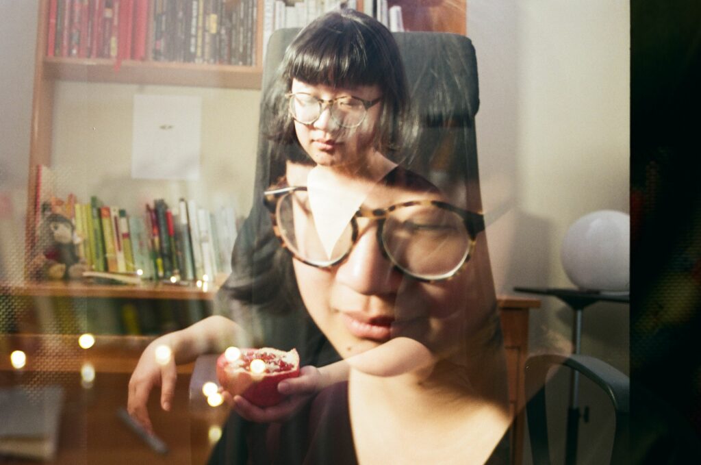 Double exposure colour photo of Jane wearing glasses, in the background image she is seated with books on shelves behind her, the foreground image is a close-up of her face