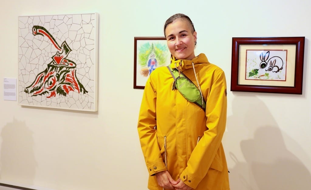 Colour photo of Kay with short dark hair, wearing a long yellow coat, standing in a gallery in front of 3 coloured images on the white wall 