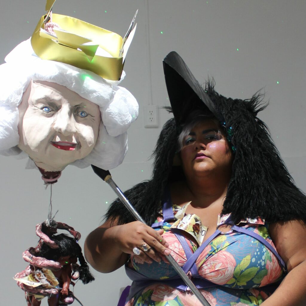 Colour photo of Raven wearing a floral dress, a large black raven headpiece with thick fur on their head, standing next to a destroyed pinata of Queen Elizabeth 