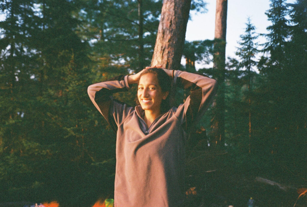 This image captures Meera's torso and head. She has her arms held up behind her head, allowing her head to rest on the palms of her hands. Her light brown hair is in a bun and her skin and eyes are brown. She is smiling with teeth visible and she is looking at the camera. She wears a grey half-zip fleece sweater. There is a large tree trunk behind her and other green pine trees in the background. 