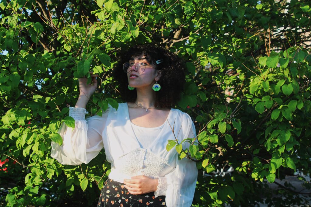 A waist-up photograph of Sama, standing in front of a large tree with green leaves, holding on to one of its branches. She is looking off to her left to something not visible in the photograph. Sama has pale skin and pink cheeks with a short head of black curls. She is wearing rounded gold-framed glasses, a white sheer top with puffed sleeves, and a black skirt with orange and white flowers on it. She is wearing beaded earrings with a green, blue, and yellow design. 